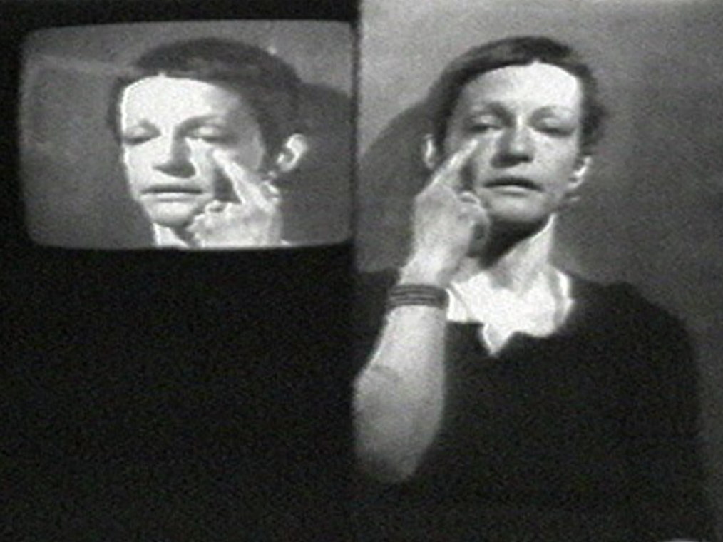 A b&w still of Joan Jonas's reflected image. She holds a pointer finger up to her eye, reflected in a CRT-style box on the left-hand side of the frame.